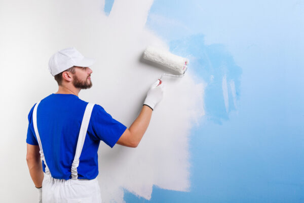 Back view of handsome young painter in white dungarees, blue t-shirt, cap and gloves painting a wall with paint roller.